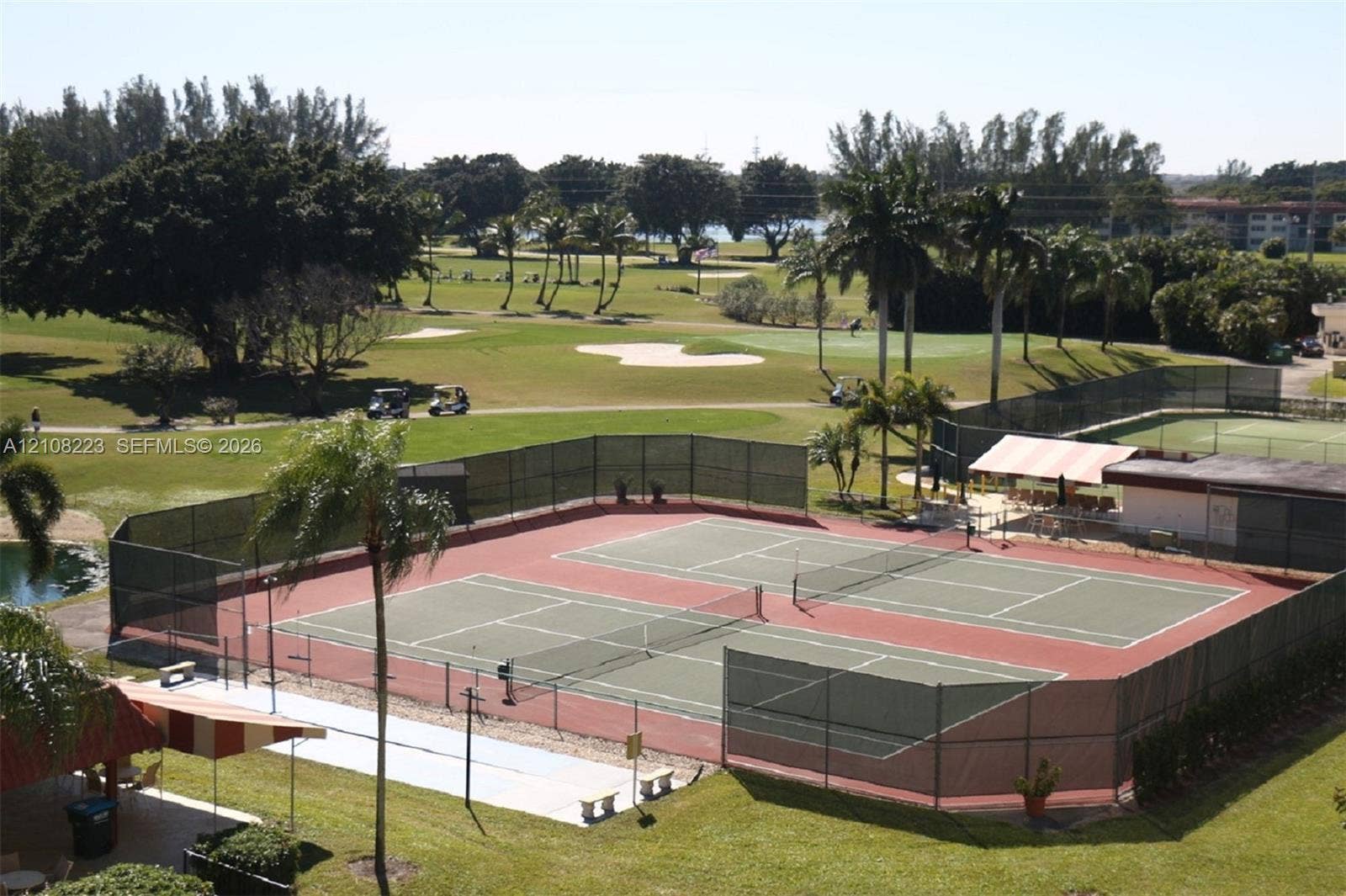 311 S Hollybrook Drive, #309, Pembroke Pines, FL 33025 Dual fenced tennis courts featuring hard-surface playing areas, accompanied by a covered pavilion, set against a backdrop of a golf course with mature landscaping and water features