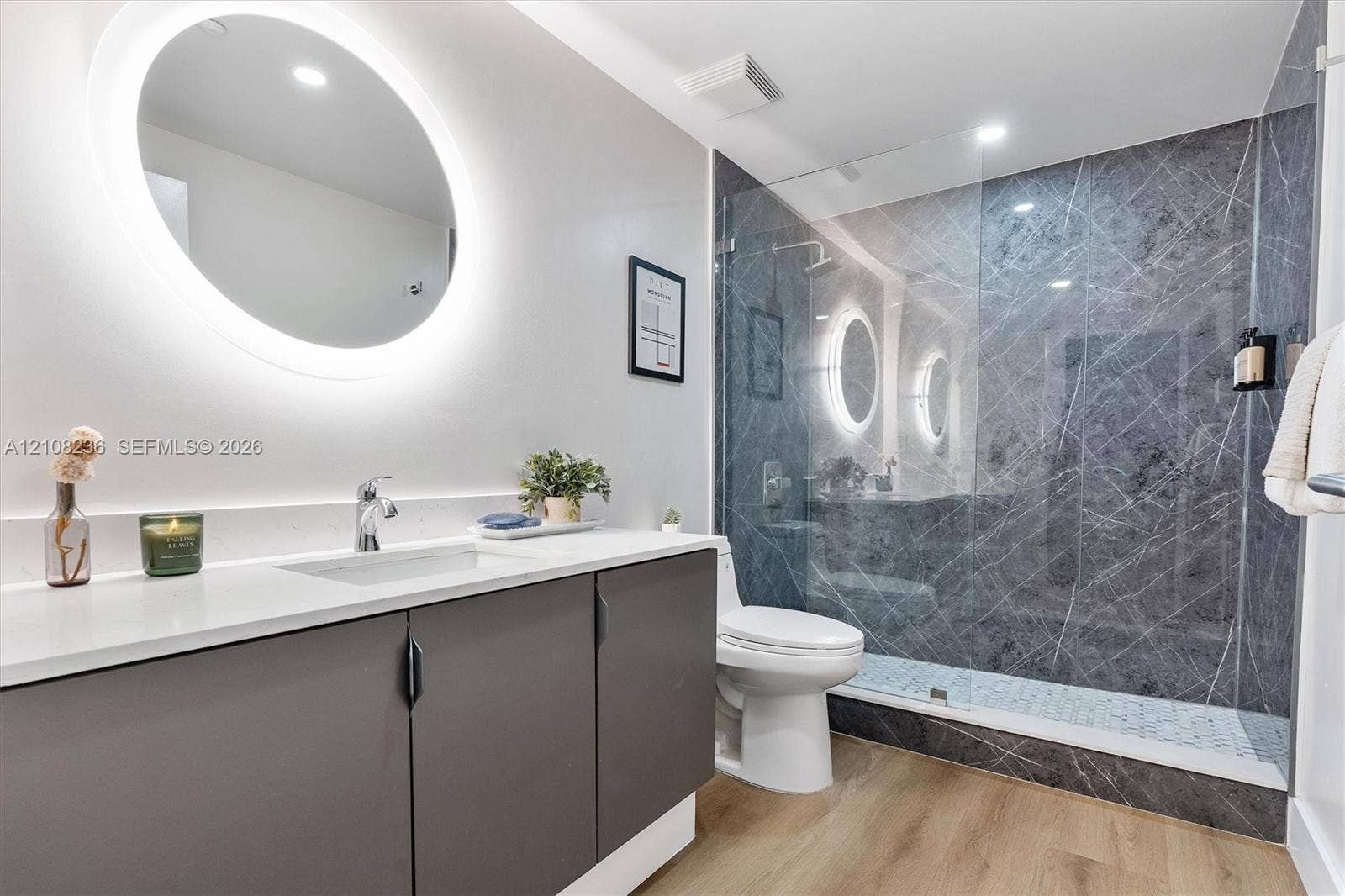 Bathroom featuring a floating vanity with a light-toned countertop, integrated oval LED-backlit mirror, wood-finish flooring, and a walk-in shower with large-format wall tile and glass enclosure