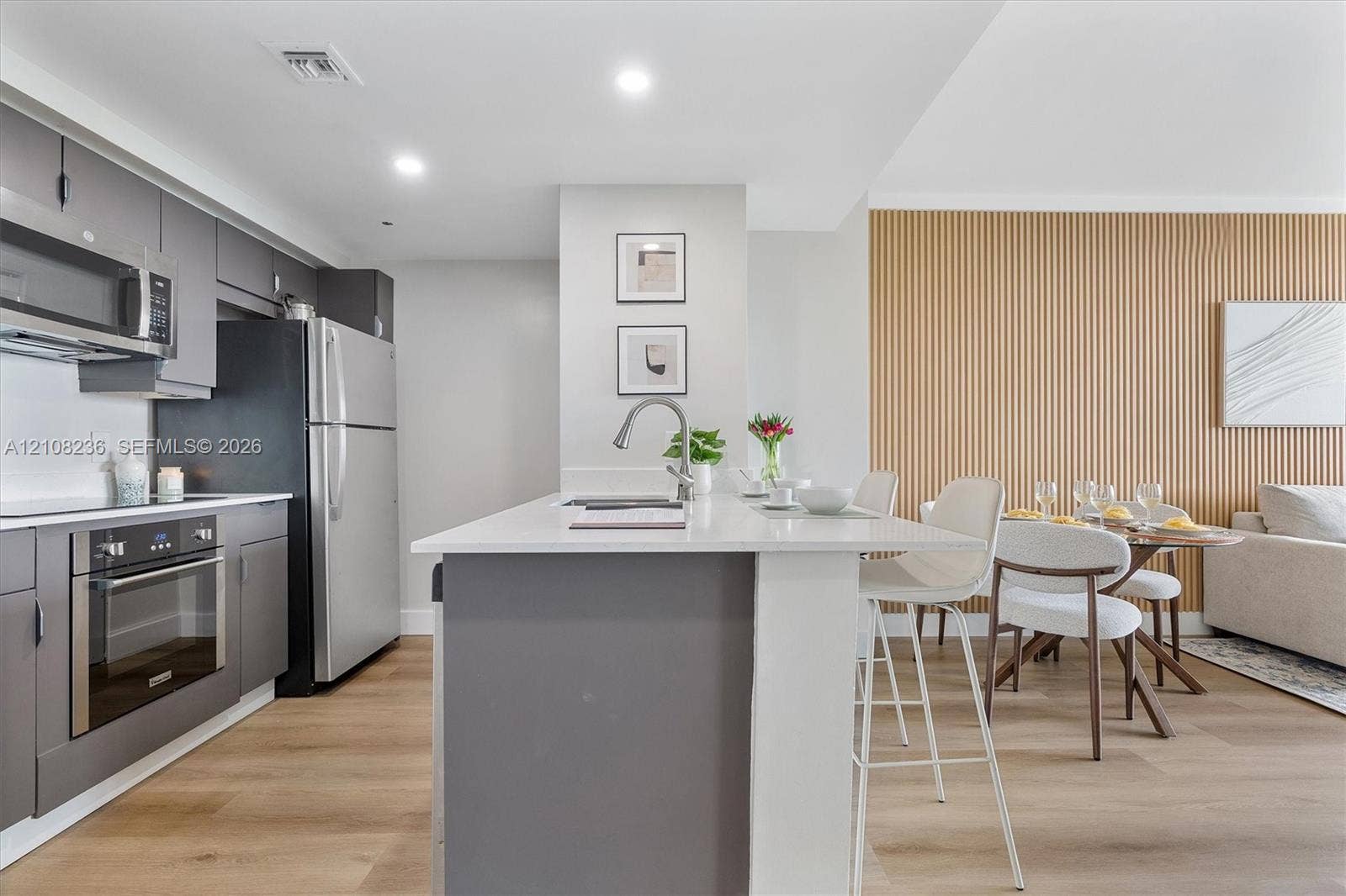 Kitchen featuring a central island with a solid-surface countertop, stainless steel appliances, flat-panel grey cabinetry, wood-finish flooring, and a decorative vertical wood-slat accent wall