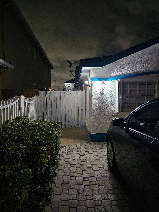 Textured stucco exterior featuring stone paver driveway, white privacy fencing, and exterior wall-mounted light fixtures