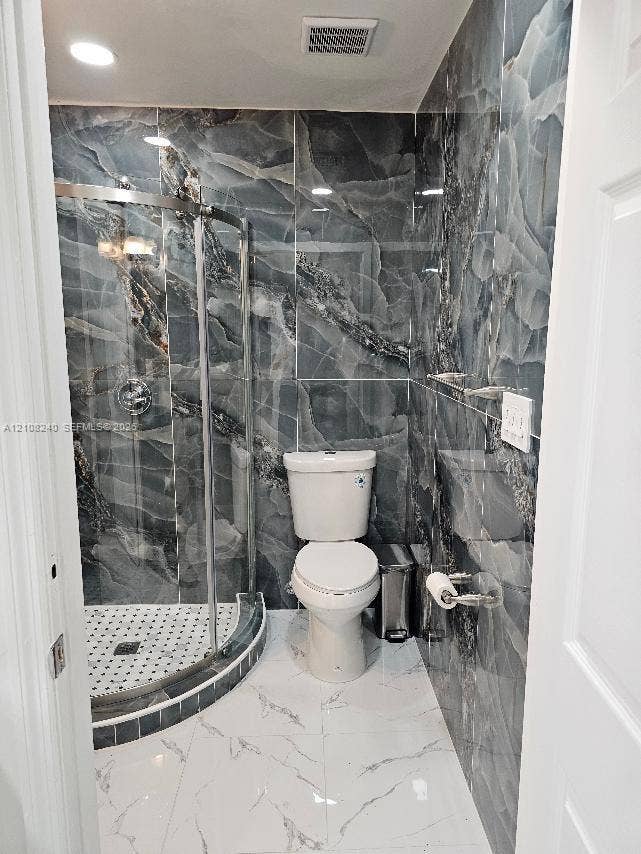 Bathroom featuring floor-to-ceiling dark-veined wall tile, a corner glass-enclosed shower with a mosaic-tile basin, and large-format white tile flooring with veining detail