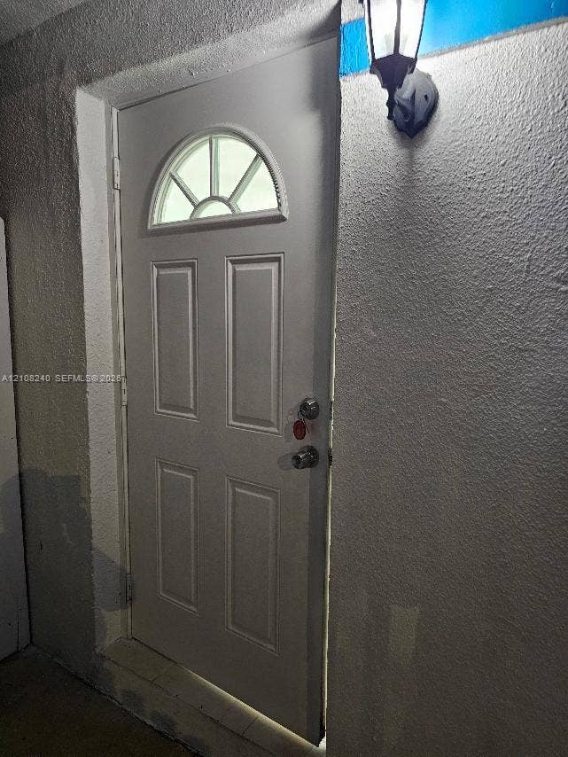 Exterior entry door featuring a semi-circular transom window, raised-panel design, and a wall-mounted lantern-style light fixture against a textured stucco exterior finish