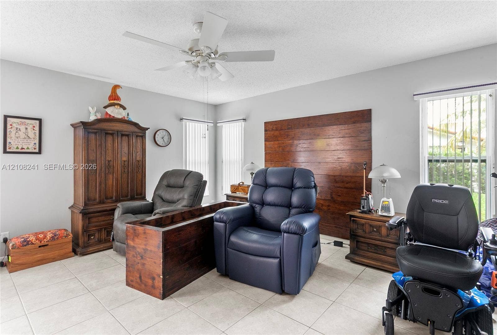 Room interior featuring light-toned tile flooring, wood-paneled accent wall, dual windows with vertical blinds, and a white ceiling fan with integrated lighting
