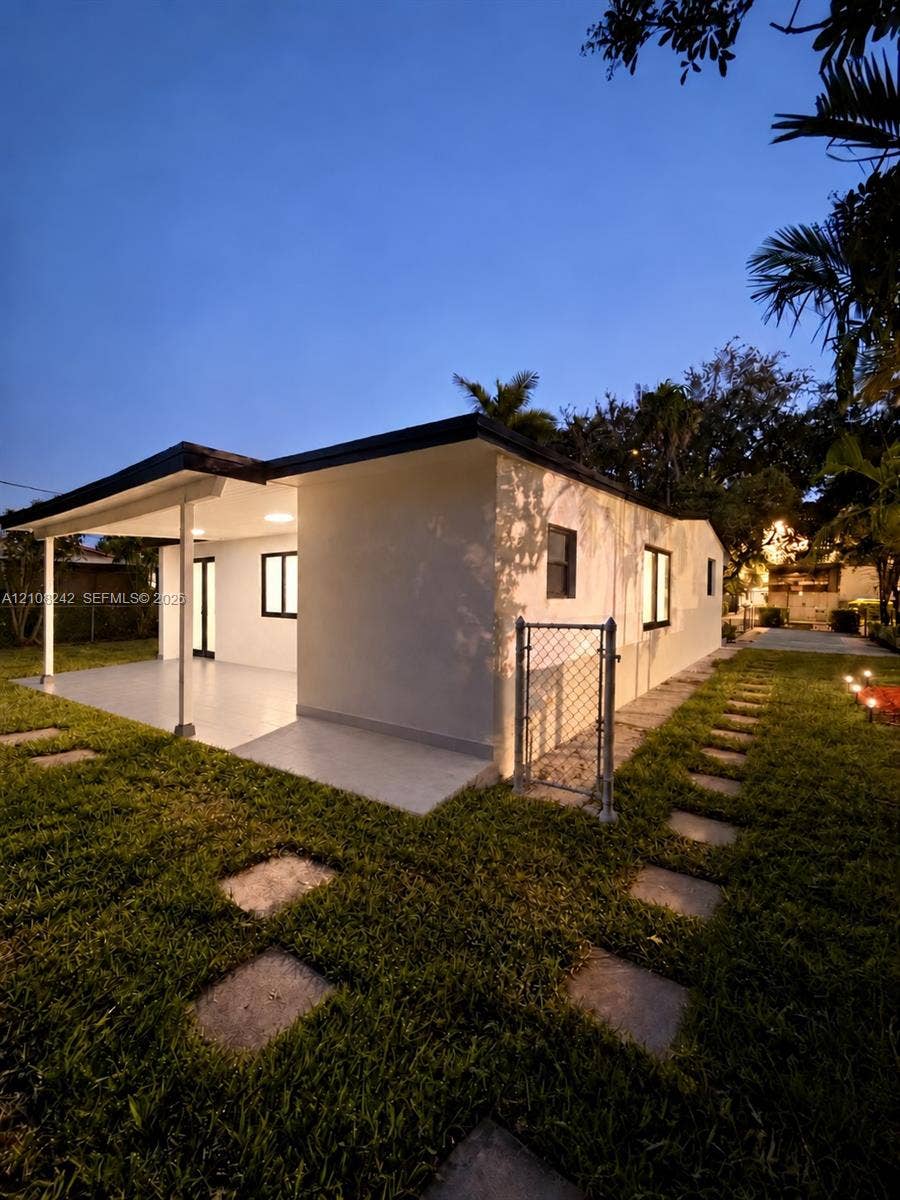 61 E 39th Street, Hialeah, FL 33013 Exterior elevation featuring a light-toned smooth stucco finish, a covered patio with tiled flooring, stone paver walkways, and a chain-link fence enclosure