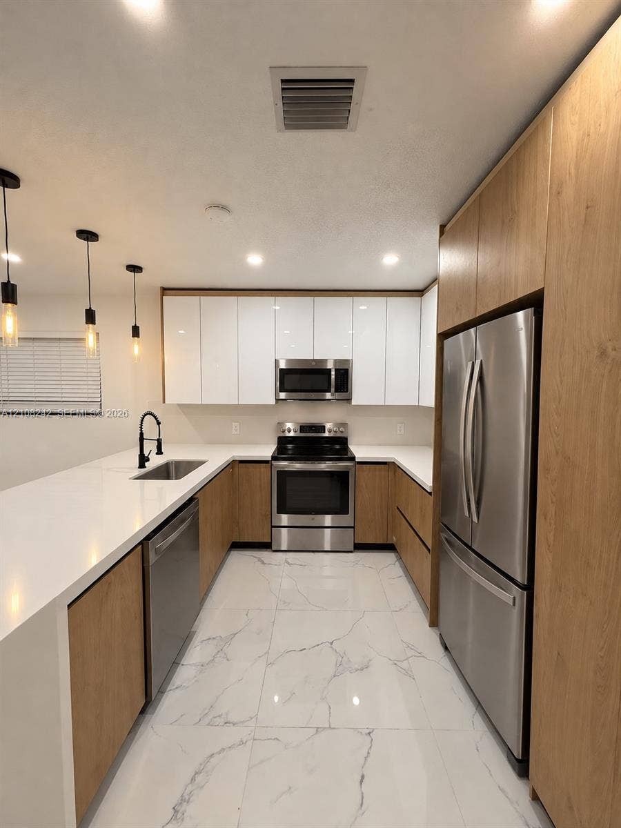 61 E 39th Street, Hialeah, FL 33013 Kitchen featuring white gloss and wood-finish cabinetry, white solid-surface countertops, large-format veined tile flooring, stainless steel appliances, and matte black pendant lighting