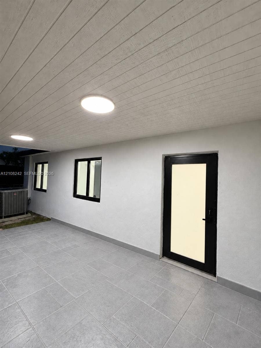 61 E 39th Street, Hialeah, FL 33013 Covered outdoor patio featuring large-format grey tile flooring, tongue-and-groove ceiling paneling, recessed lighting, and a modern glass-panel door with a black frame