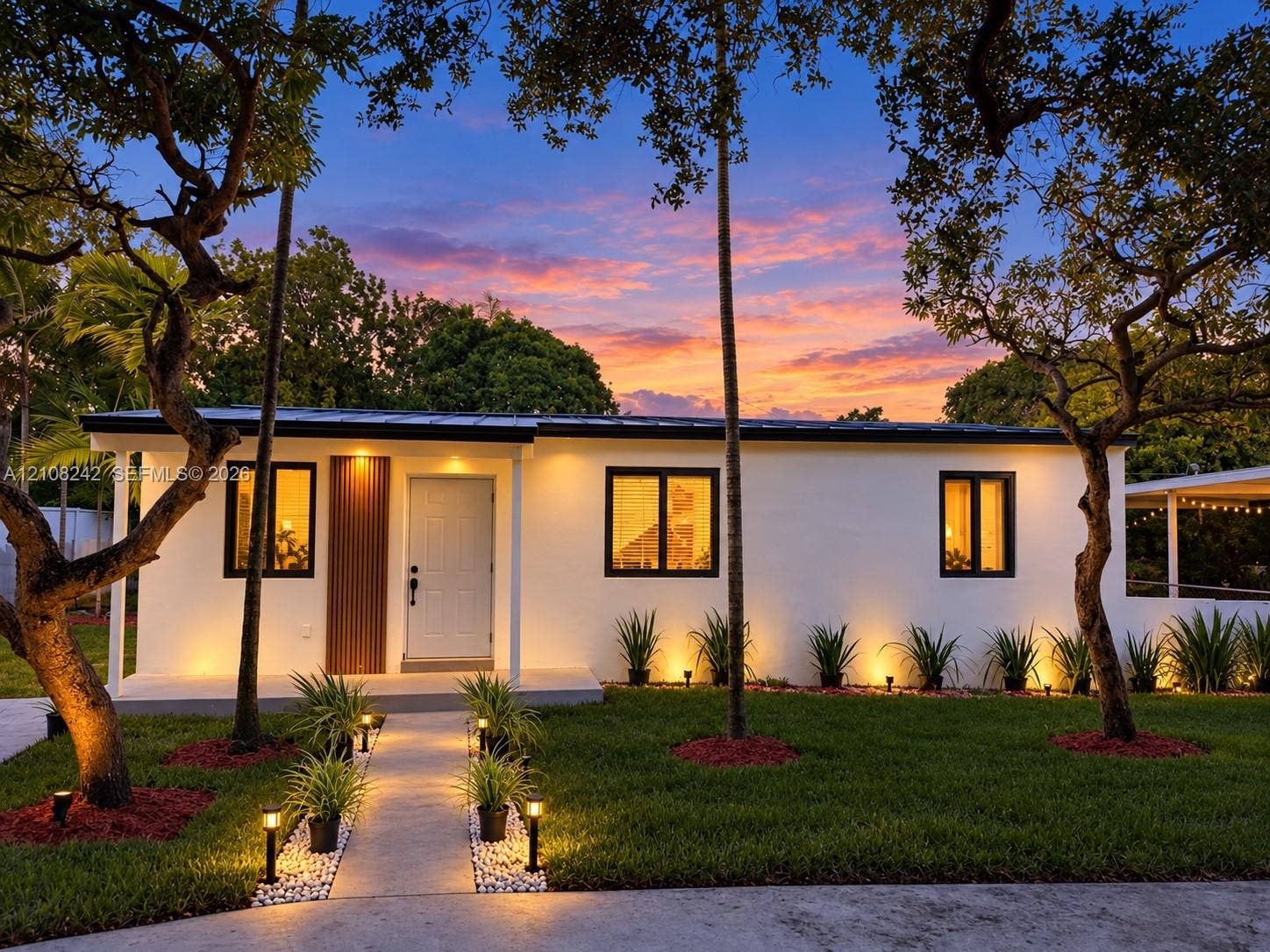 61 E 39th Street, Hialeah, FL 33013 Single-story residence featuring a smooth stucco exterior, dark-trimmed windows, a metal roof, and a vertical wood-slat accent wall at the entryway, complemented by a landscaped front yard with a concrete walkway