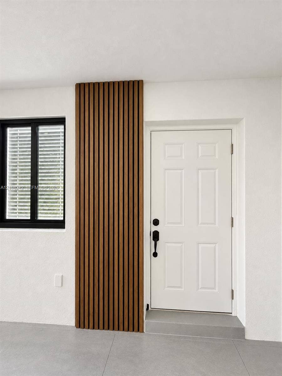 61 E 39th Street, Hialeah, FL 33013 Vertical wood-slat accent wall, six-panel entry door with matte black hardware, black-framed window, and neutral large-format floor tiling