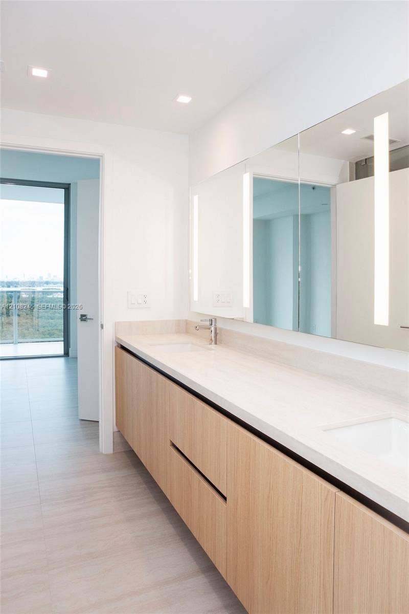 Floating vanity featuring a wood-finish cabinet, dual undermount sinks, stone-finish countertop, full-width mirror with integrated vertical lighting, and recessed ceiling lighting