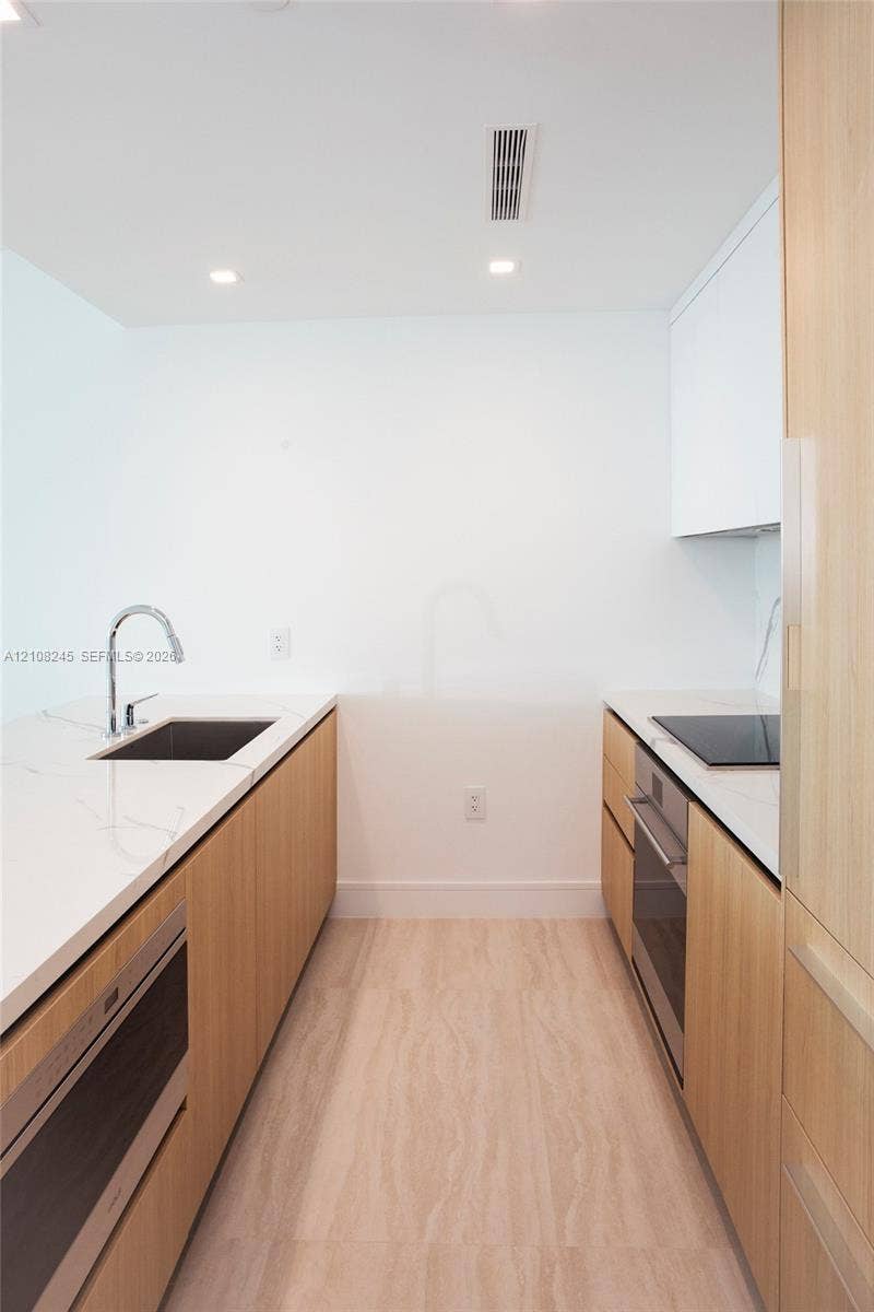 Kitchen galley featuring light wood-finish cabinetry, white stone-finish countertops, an undermount sink with a gooseneck faucet, a built-in oven, and a cooktop