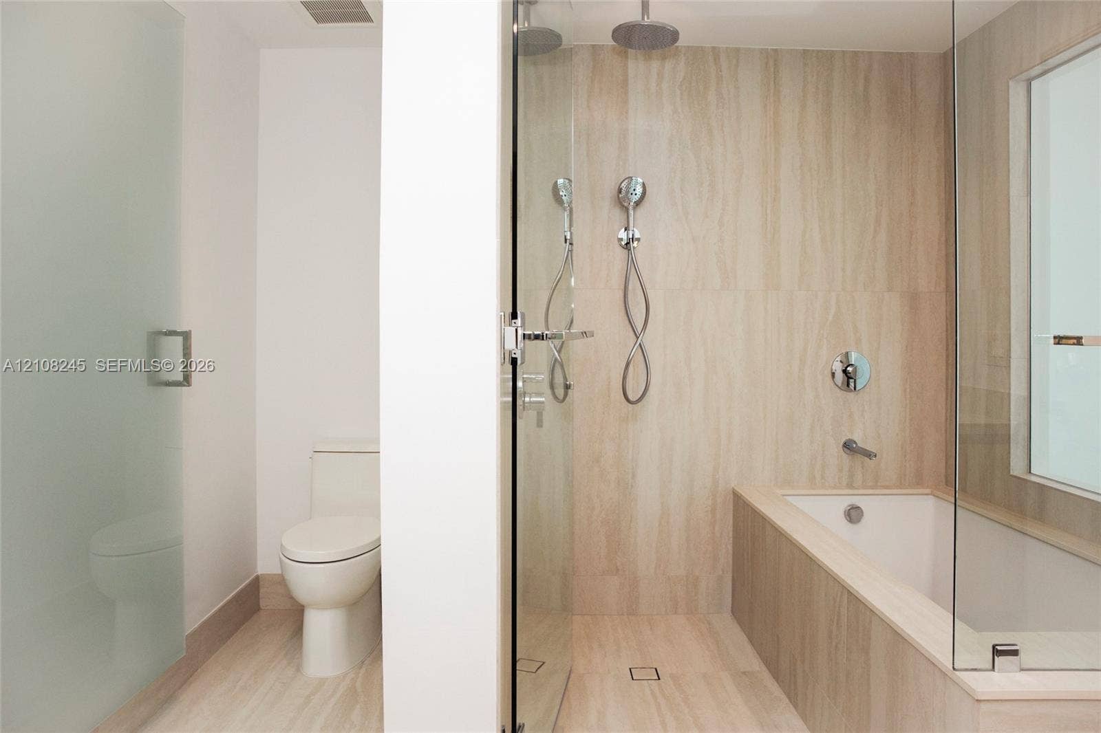Bathroom featuring stone-finish wall and floor tiling, glass-enclosed shower with rainfall and handheld showerheads, deep soaking tub, and enclosed toilet area