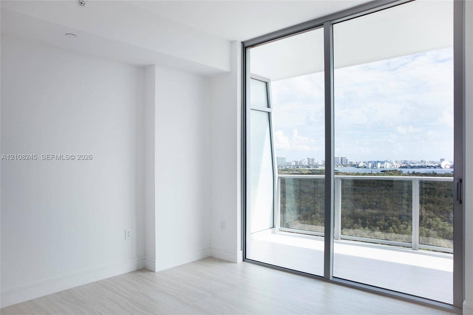 Floor-to-ceiling glass sliding doors, wood-finish flooring, and a private balcony featuring a glass railing and urban skyline scenery