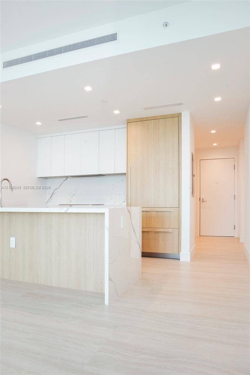 Kitchen featuring waterfall-edge stone-finish island, light wood-finish cabinetry, white flat-panel upper cabinets, integrated appliances, and recessed ceiling lighting