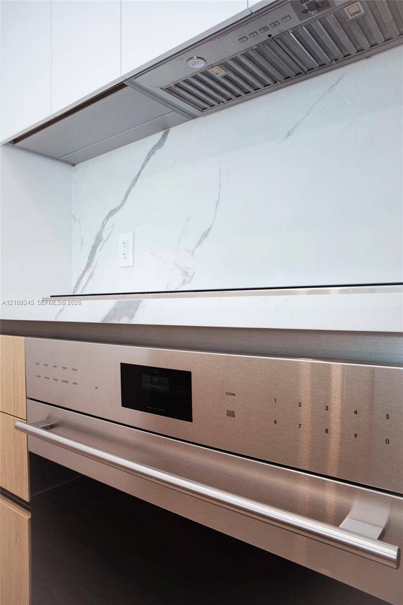 Sleek stainless steel wall oven, stone-finish backsplash with integrated hood range, and wood-finish cabinetry