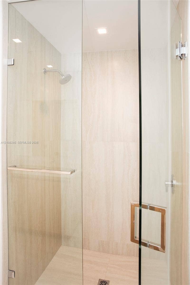 Walk-in shower enclosure featuring floor-to-ceiling neutral stone-finish tile, frameless glass paneling, polished chrome hardware, and an integrated wall-mounted showerhead