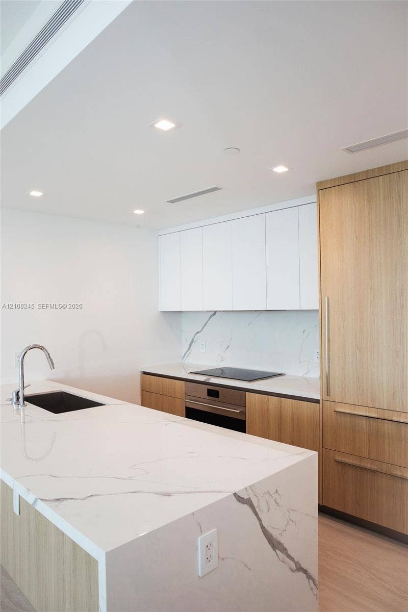 Kitchen featuring a central island with a stone countertop and waterfall edge, custom wood-finish cabinetry, white upper cabinets, integrated stainless steel appliances, and a stone slab backsplash