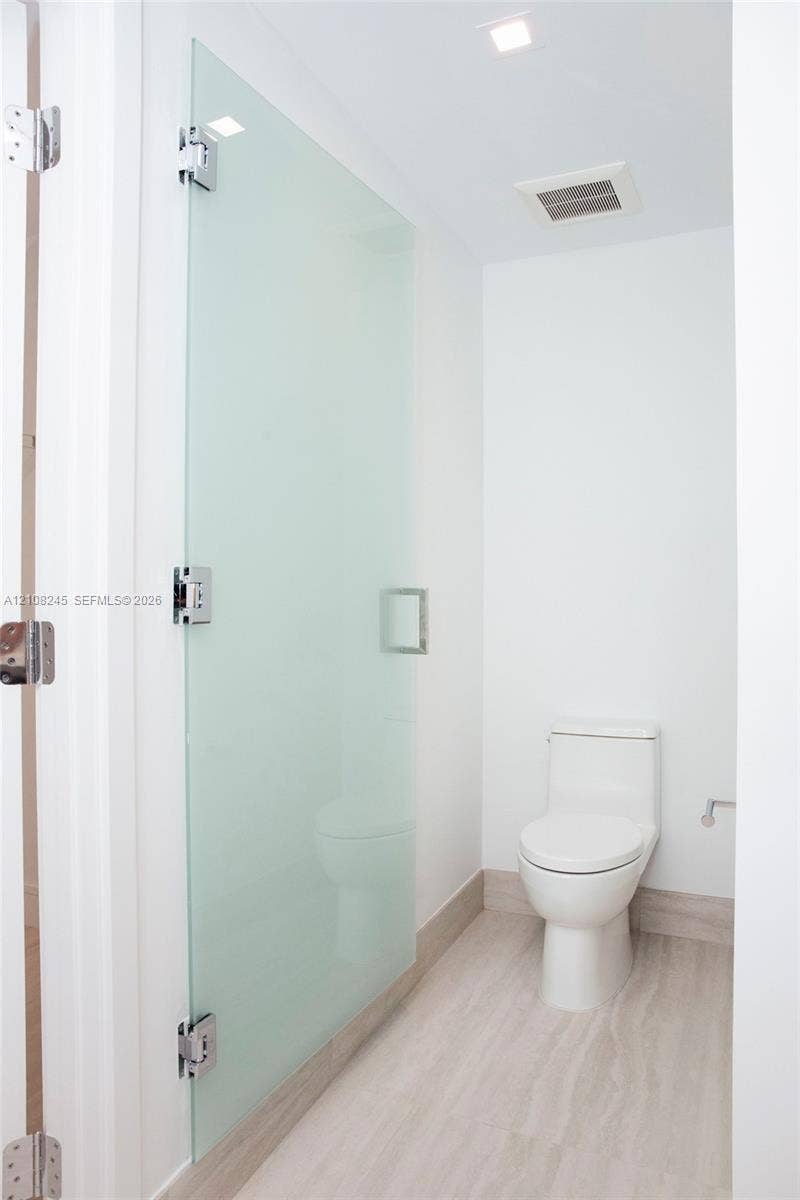Frosted glass panel with polished chrome hardware, white porcelain elongated toilet, and neutral-tone wood-finish flooring