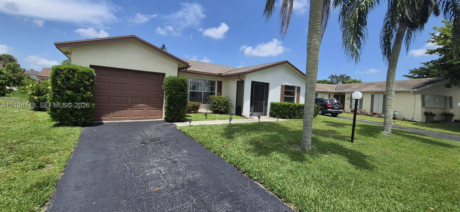 4490 Pine Garden Lane, Lake Worth, FL 33467 Single-story residence featuring a light-colored stucco exterior, attached garage with a solid-panel door, paved driveway, screen-enclosed entryway, and mature palm trees