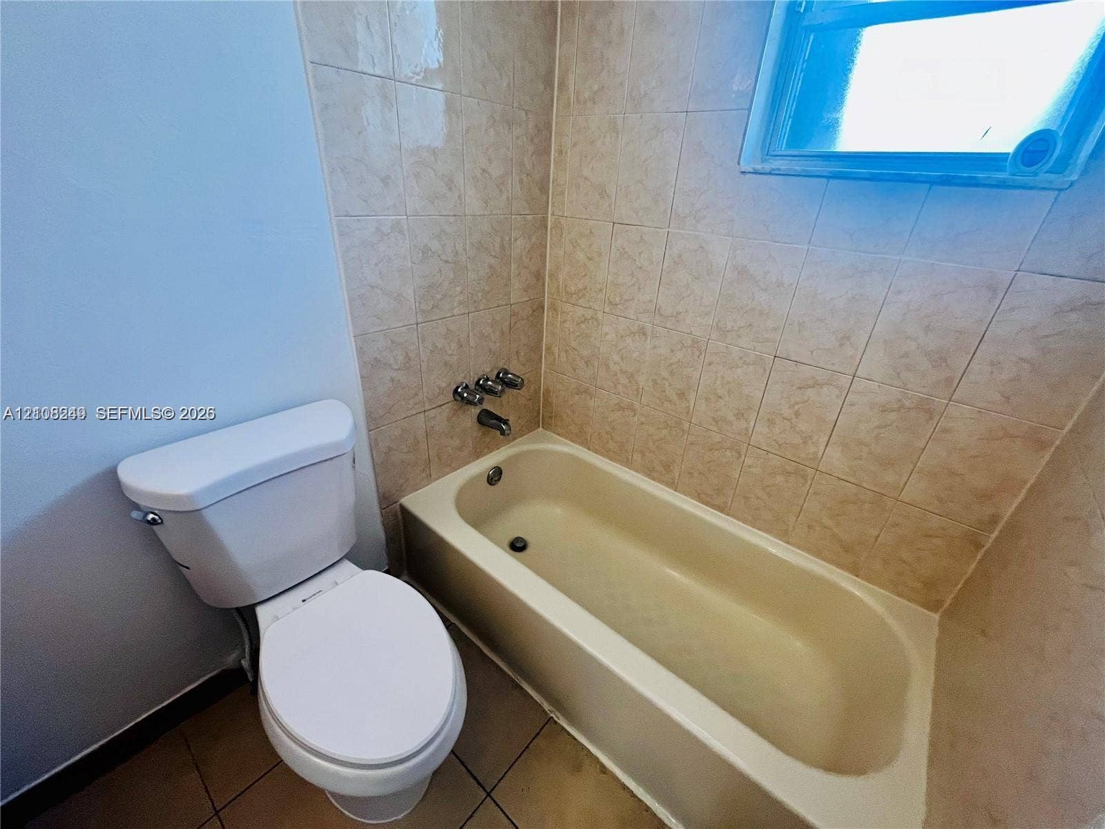 Bathroom featuring a porcelain tub, neutral ceramic wall tile, a window, and a standard-height toilet
