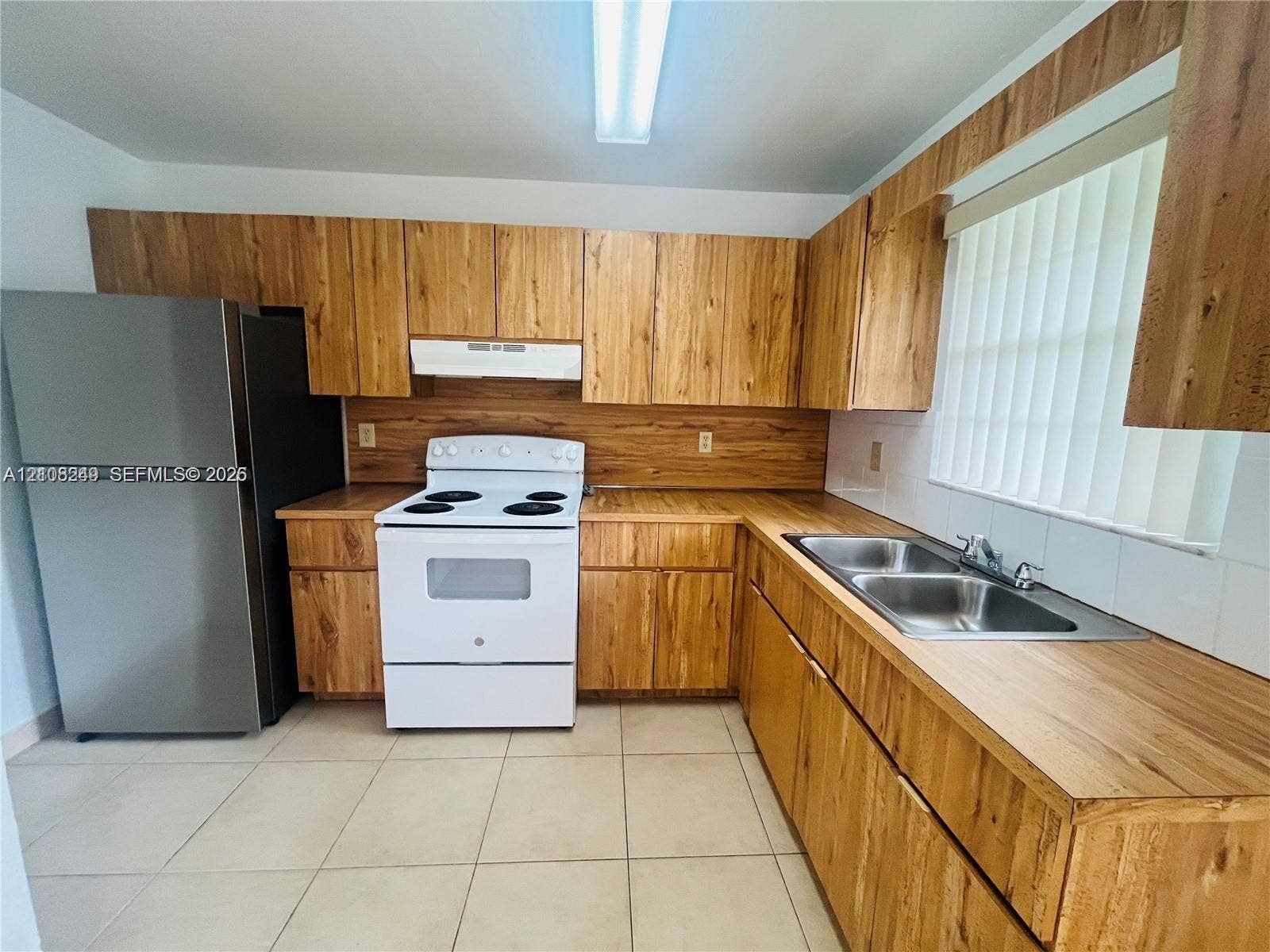 Kitchen featuring wood-finish cabinetry, wood-finish countertops, tile flooring, a dual-basin stainless steel sink, and stainless steel refrigerator