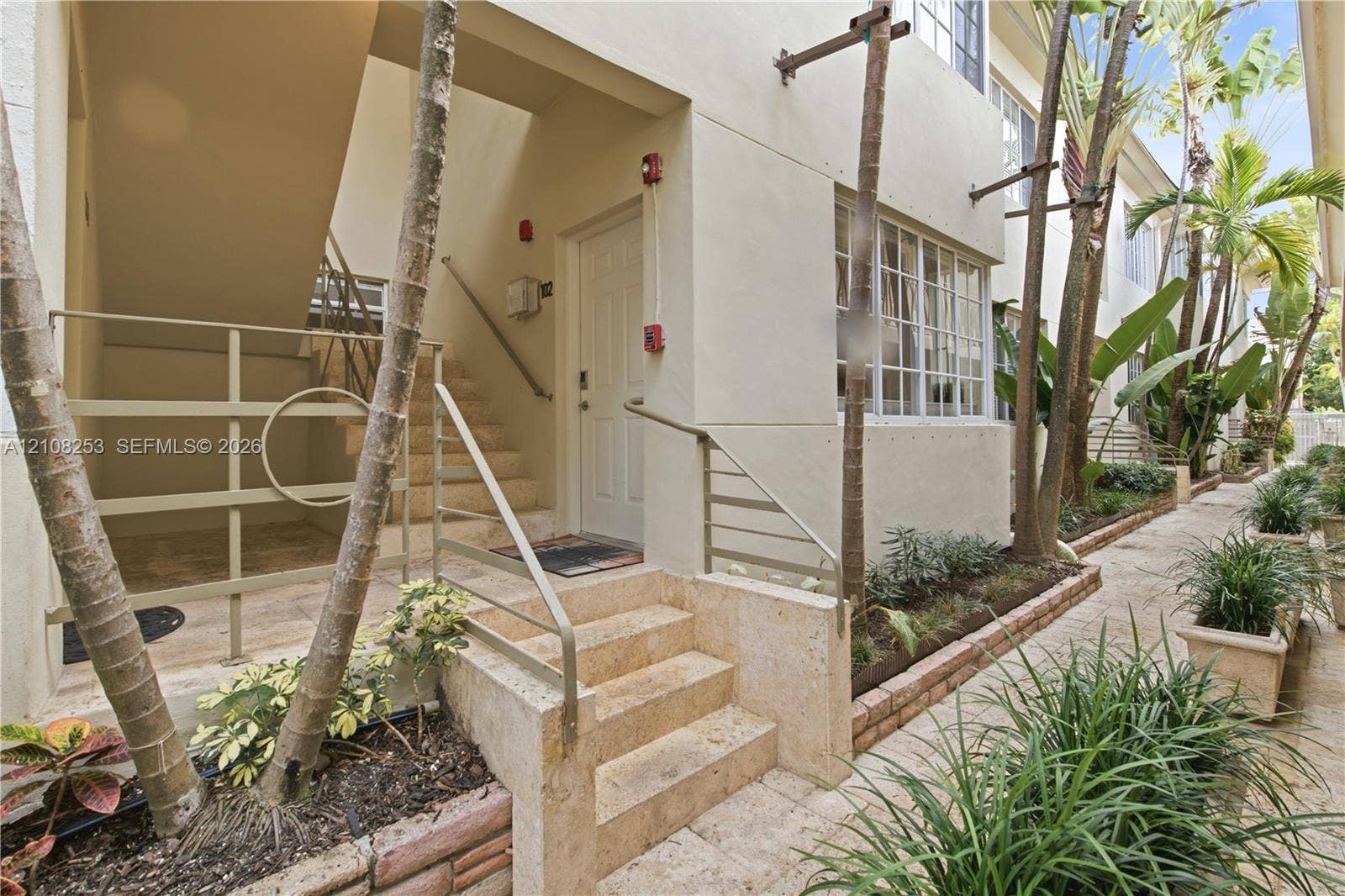 1330 Drexel Avenue, #102, Miami Beach, FL 33139 Exterior walkway featuring natural stone pavers, concrete stairs, metal handrails, and built-in landscape planters with mature palms and shrubbery