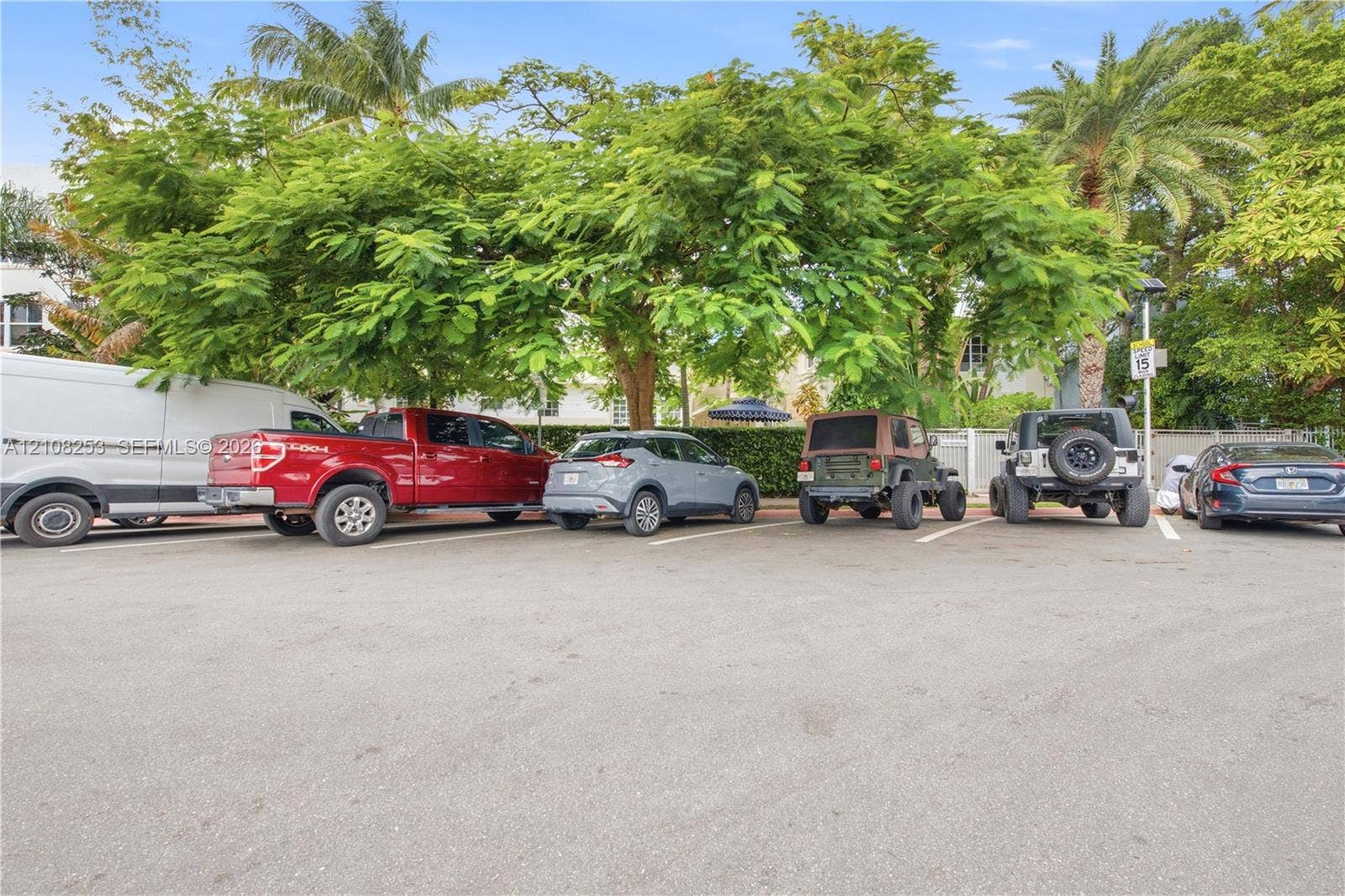 1330 Drexel Avenue, #102, Miami Beach, FL 33139 Asphalt parking area featuring marked spaces, perimeter privacy hedging, and mature canopy trees