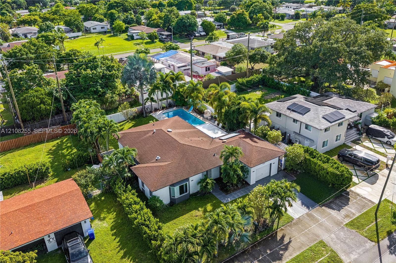 1650 NE 171st Street, North Miami Beach, North Miami Beach, FL 33162 Aerial perspective of a single-story residence featuring a shingled hip roof, a private rectangular swimming pool, integrated solar panels, and manicured landscaping with mature palm trees