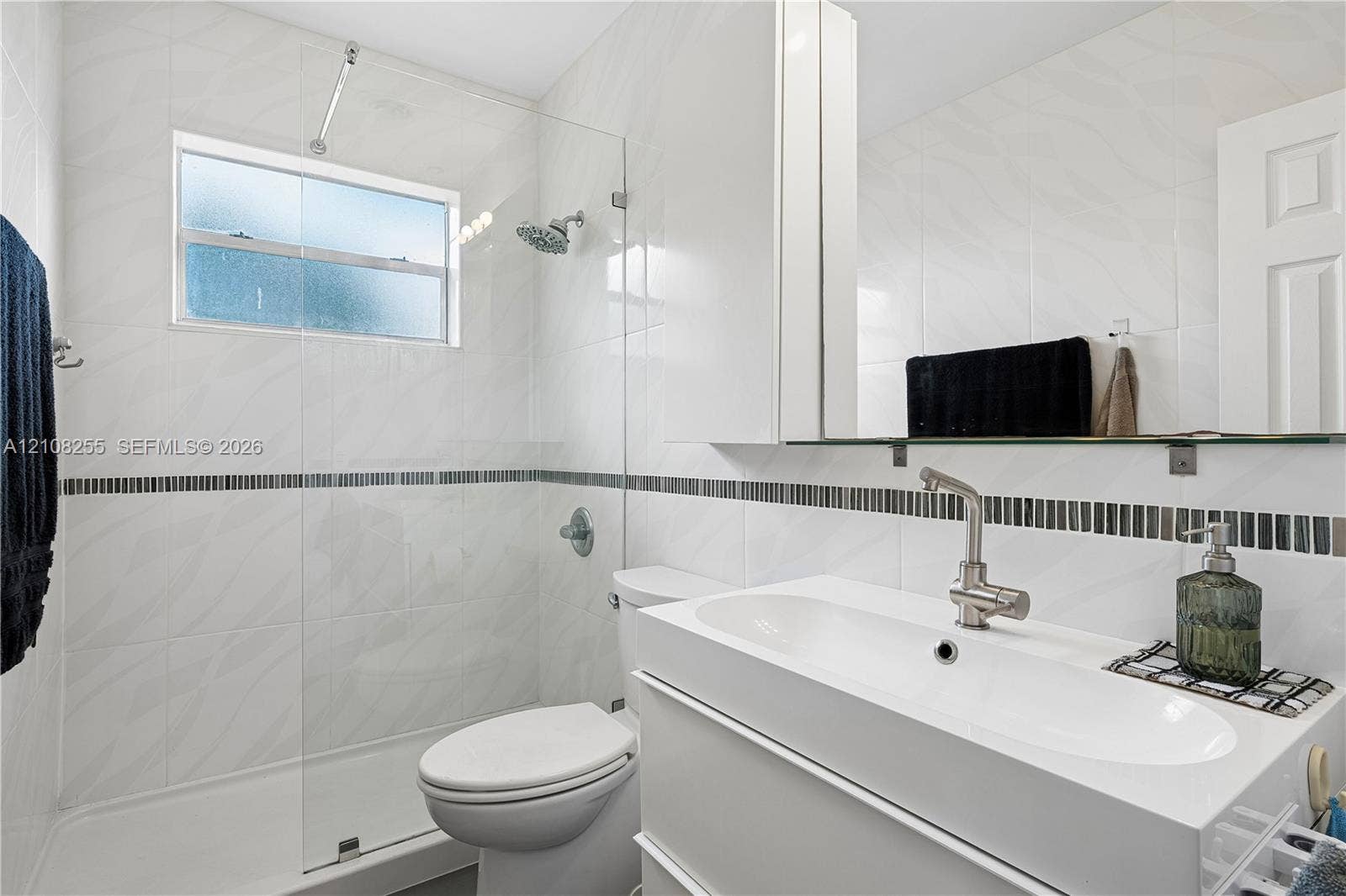 1650 NE 171st Street, North Miami Beach, North Miami Beach, FL 33162 Bathroom featuring white wall tile with a mosaic accent border, a glass-enclosed shower, a floating vanity with an integrated sink, and brushed-nickel hardware