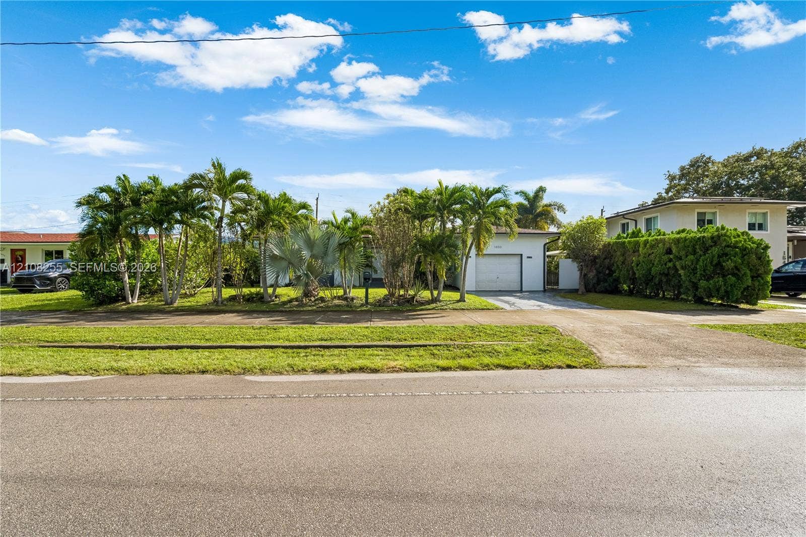1650 NE 171st Street, North Miami Beach, North Miami Beach, FL 33162 Exterior of a single-story residence featuring a concrete driveway, attached garage, manicured lawn, and mature palm landscaping