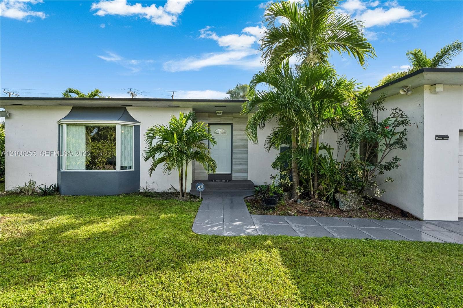 1650 NE 171st Street, North Miami Beach, North Miami Beach, FL 33162 Single-story exterior featuring white stucco finish, front bay window, stone-accented entryway, gray concrete walkway, and established palm landscaping