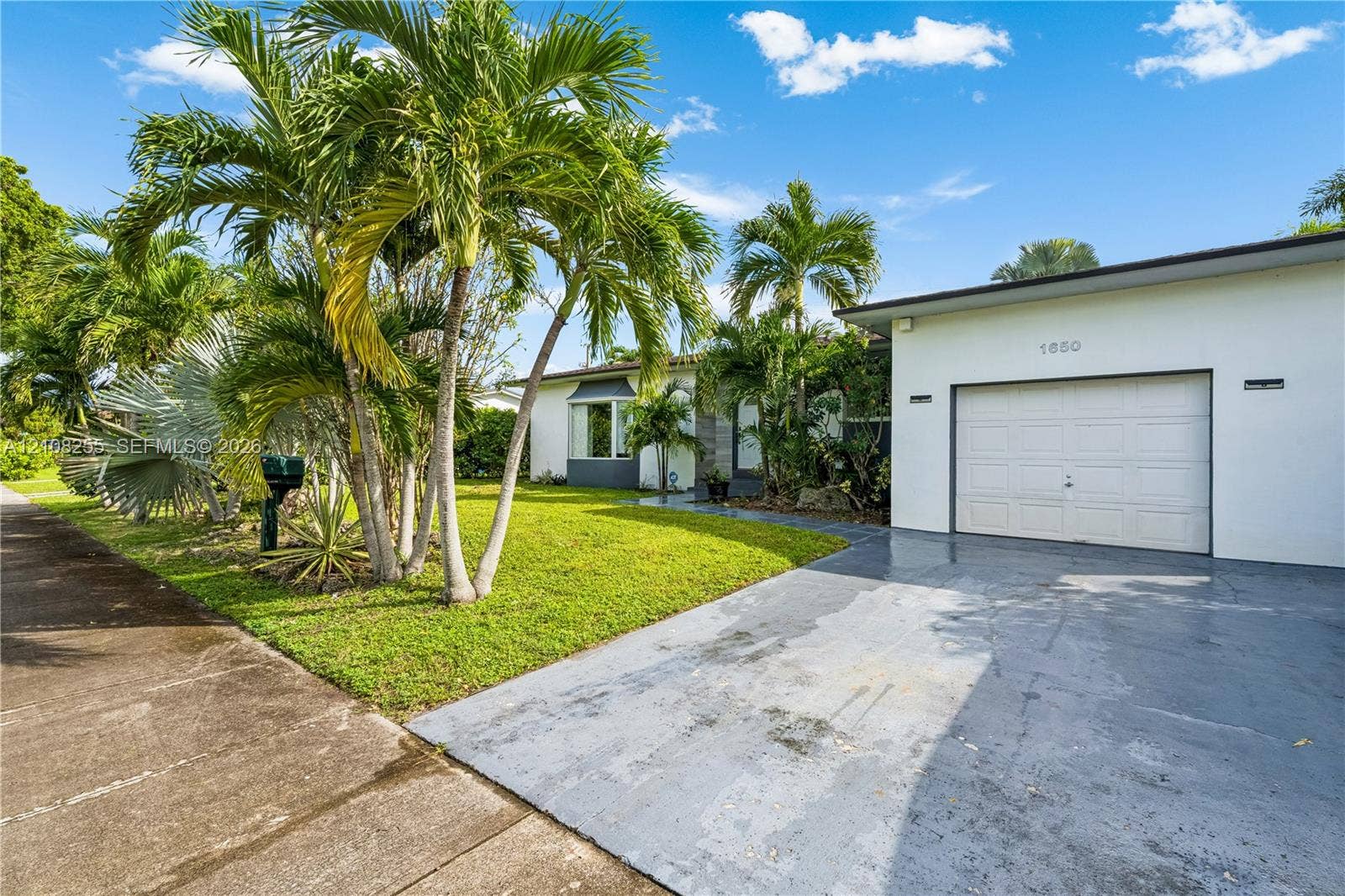 1650 NE 171st Street, North Miami Beach, North Miami Beach, FL 33162 Single-story exterior featuring white stucco siding, a paved driveway, a one-car garage, and established palm landscaping