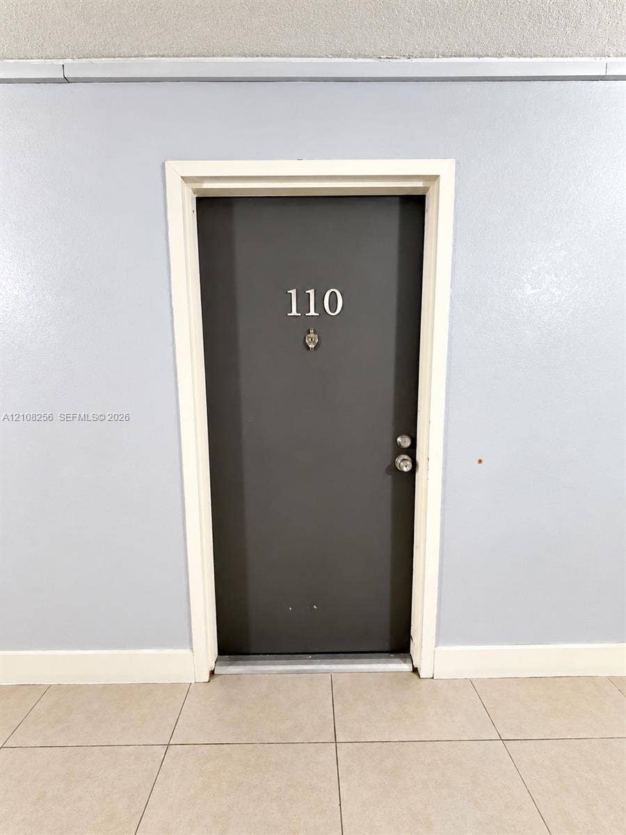 9350 Fontainebleau Boulevard, #110, Miami, Miami, FL 33172 Dark-toned entry door featuring a polished metal door handle, deadbolt lock, and metal kick plate, framed by white trim against light-toned walls and tile flooring