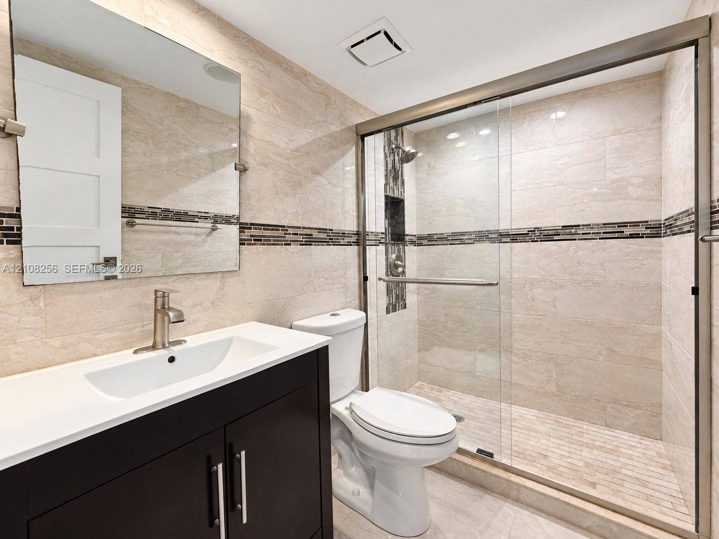 9350 Fontainebleau Boulevard, #110, Miami, Miami, FL 33172 Bathroom featuring a dark wood-finish vanity with a white countertop, walk-in shower with sliding glass doors, neutral wall tiling with a mosaic accent border, and a frameless mirror