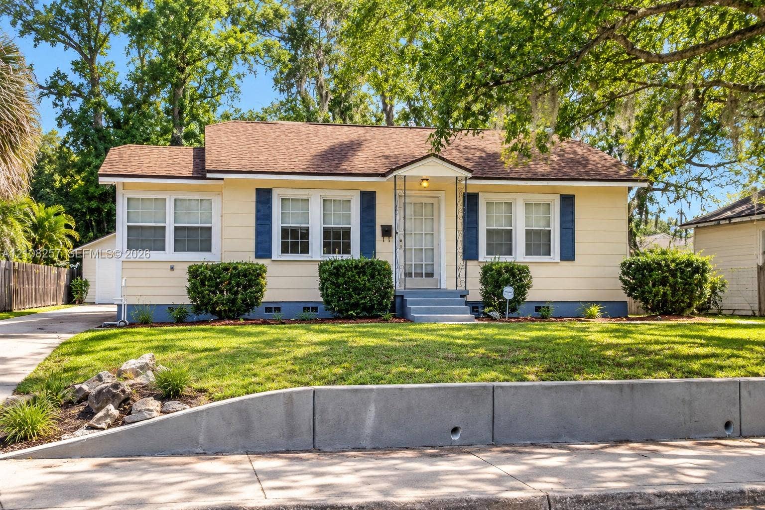 Single-story exterior featuring light-toned horizontal siding, decorative shutters, asphalt shingle roofing, and a concrete retaining wall