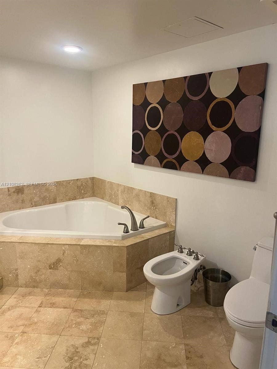 Corner soaking tub with natural stone surround, stone tile flooring, bidet, and two-piece toilet