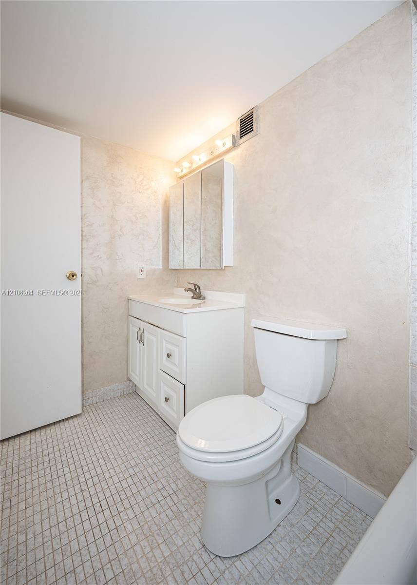 Bathroom featuring white vanity cabinetry with integrated countertop, tri-fold medicine cabinet, wall-mounted light fixture, and ceramic tile flooring