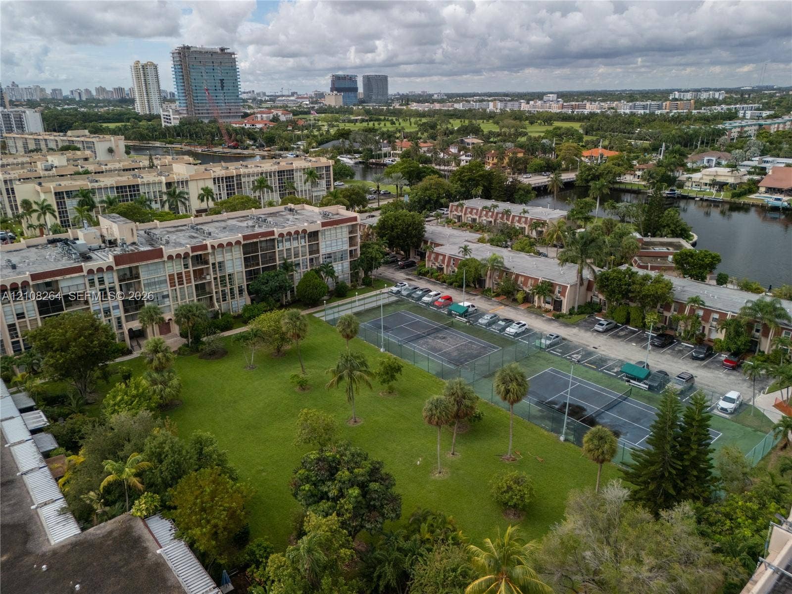 Aerial perspective of community grounds featuring two tennis courts, open green space, mature palm trees, and canal frontage