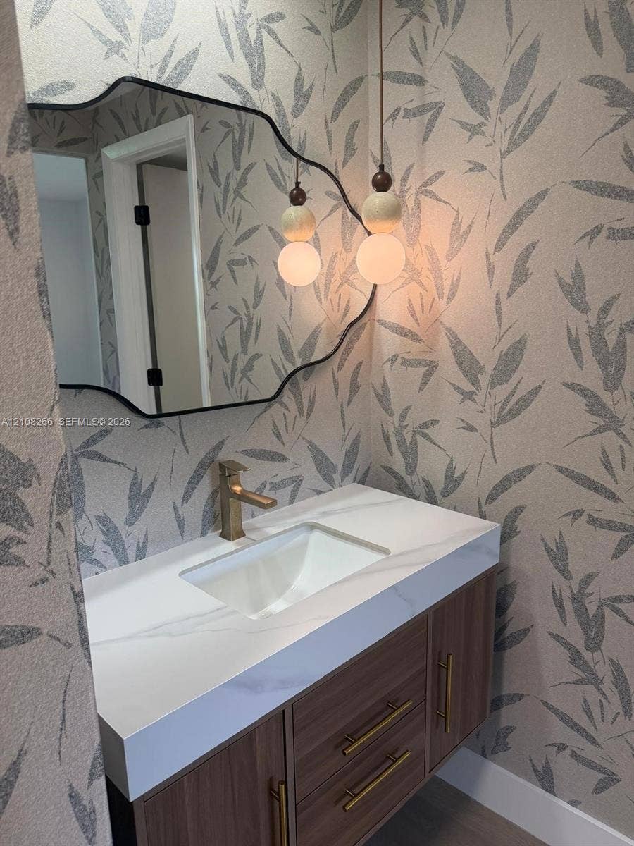 9721 Sunrise Lakes Boulevard, #108, Sunrise, FL 33322 Floating vanity with wood-finish cabinetry, white countertop, integrated basin, brushed gold-tone hardware, and sculptural pendant lighting set against patterned wall covering with a black-framed accent mirror
