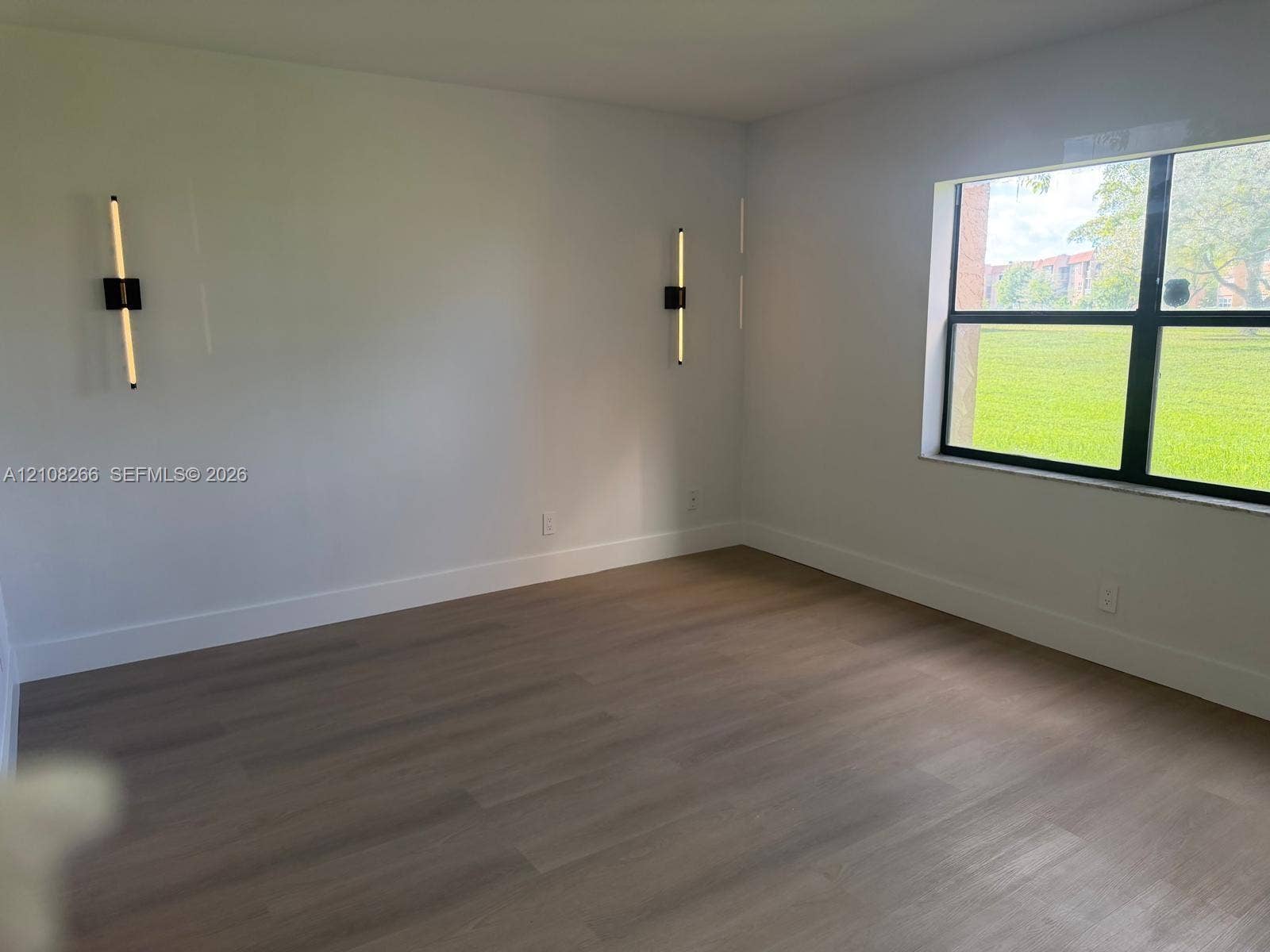 9721 Sunrise Lakes Boulevard, #108, Sunrise, FL 33322 Neutral-toned wood-finish flooring, two modern vertical wall-mounted fixtures, and a large window with black framing