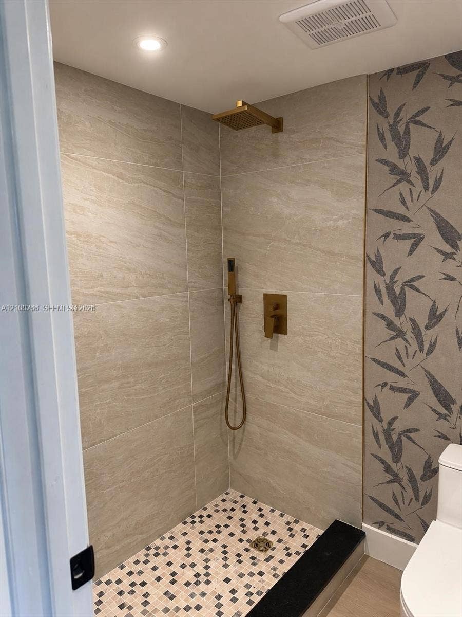 9721 Sunrise Lakes Boulevard, #108, Sunrise, FL 33322 Walk-in shower featuring large-format stone-finish tile surround, mosaic floor tiling, gold-tone rainfall showerhead, and matching hand-held sprayer