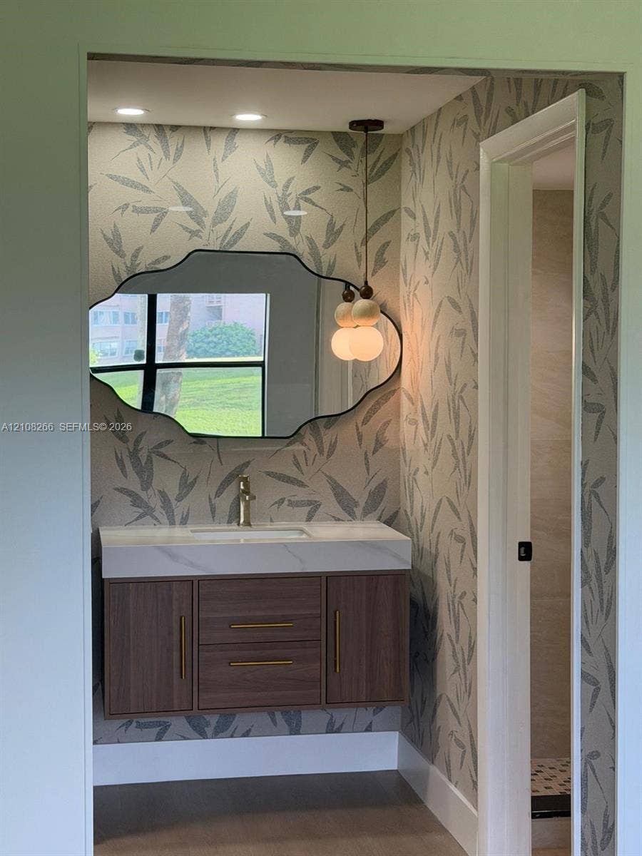 9721 Sunrise Lakes Boulevard, #108, Sunrise, FL 33322 Floating wood-finish vanity with a quartz countertop, integrated basin, brushed brass hardware, scalloped-edge wall mirror, and dual-globe pendant lighting