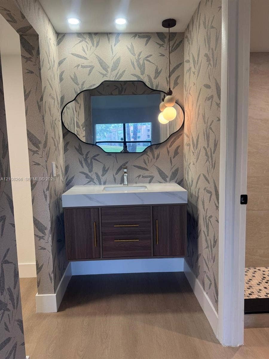9721 Sunrise Lakes Boulevard, #108, Sunrise, FL 33322 Floating wood-finish vanity with a rectangular sink, quartz countertop, custom-shaped wall mirror, and modern pendant lighting