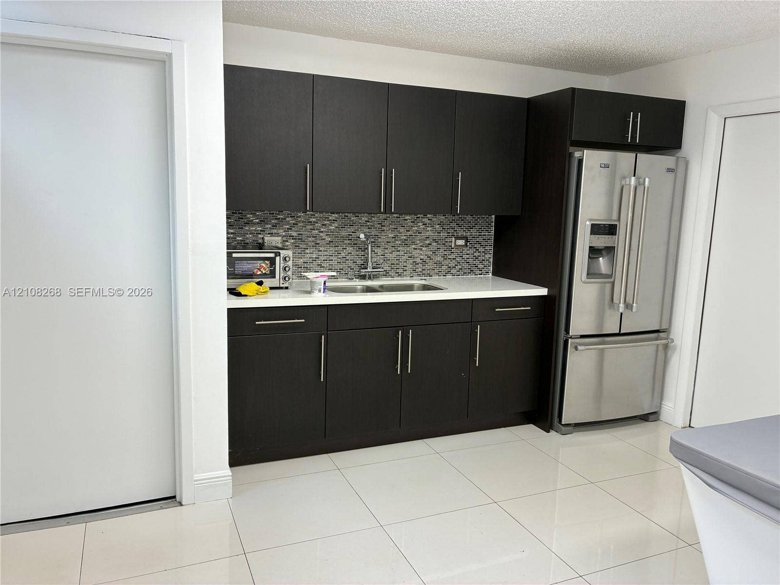 Dark espresso cabinetry, white solid-surface countertops, mosaic tile backsplash, stainless steel refrigerator, and ceramic tile flooring