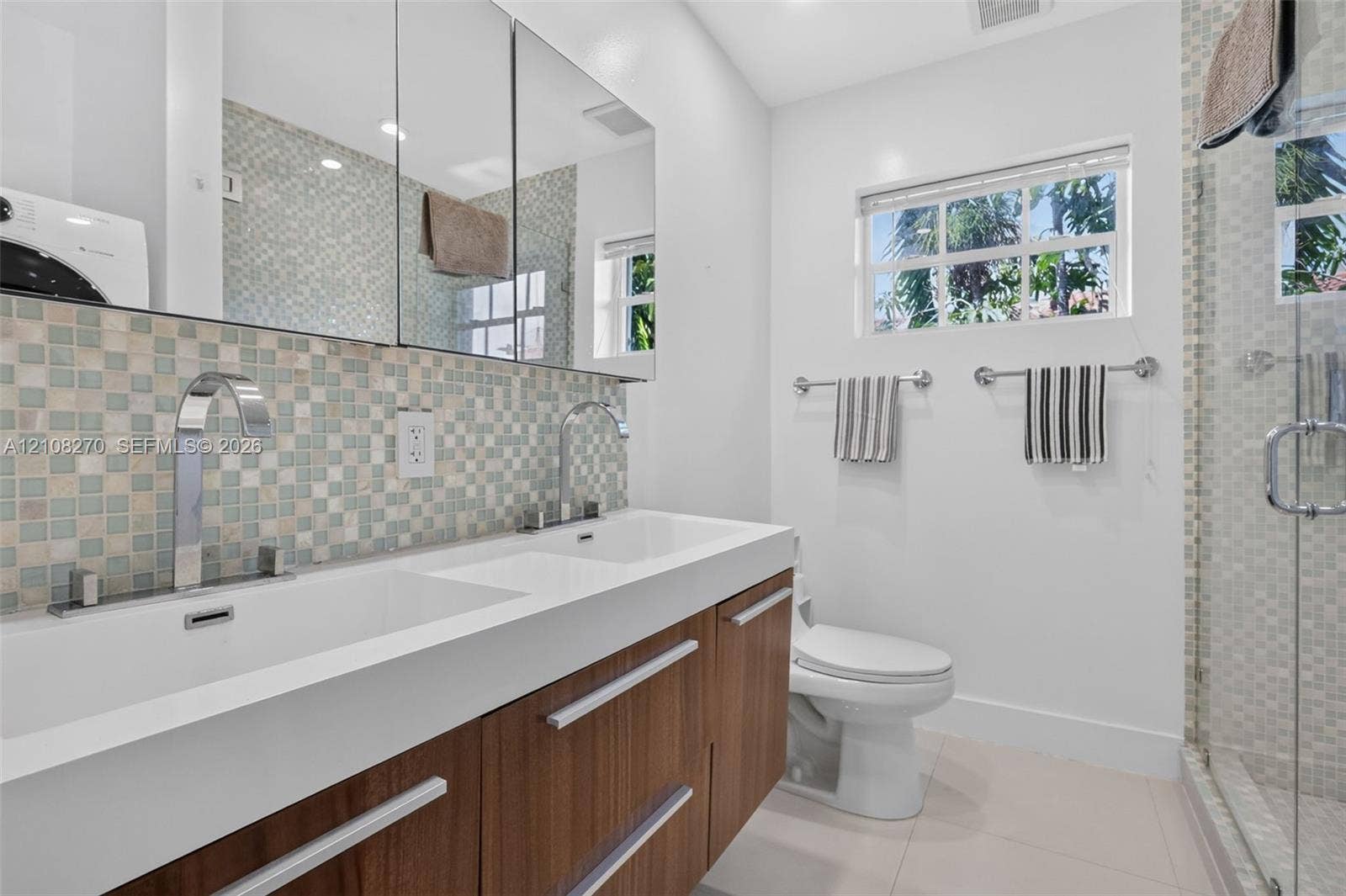 1125 99th Street, #12, Bay Harbor Islands, FL 33154 Dual-sink floating vanity with wood-finish cabinetry, mosaic tile backsplash, mirrored medicine cabinets, and glass-enclosed shower
