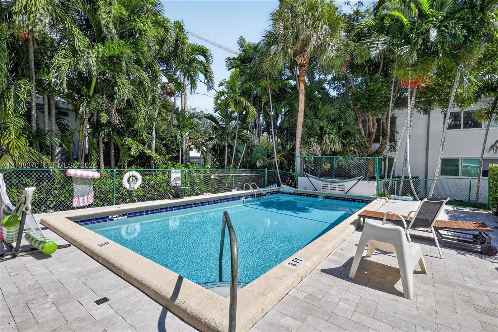 1125 99th Street, #12, Bay Harbor Islands, FL 33154 Rectangular swimming pool featuring a tile border and metal grab rails, surrounded by a paved patio and enclosed by perimeter fencing with mature palm trees