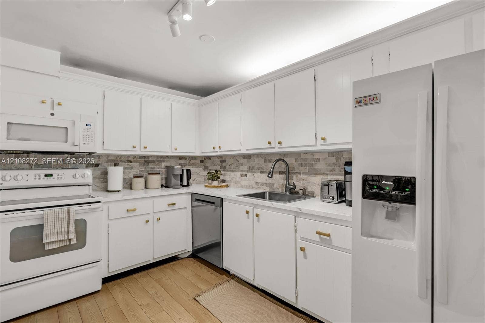 Kitchen featuring white cabinetry with gold-tone hardware, stone-style tile backsplash, white countertops, stainless steel undermount sink with a gooseneck faucet, and wood-finish flooring