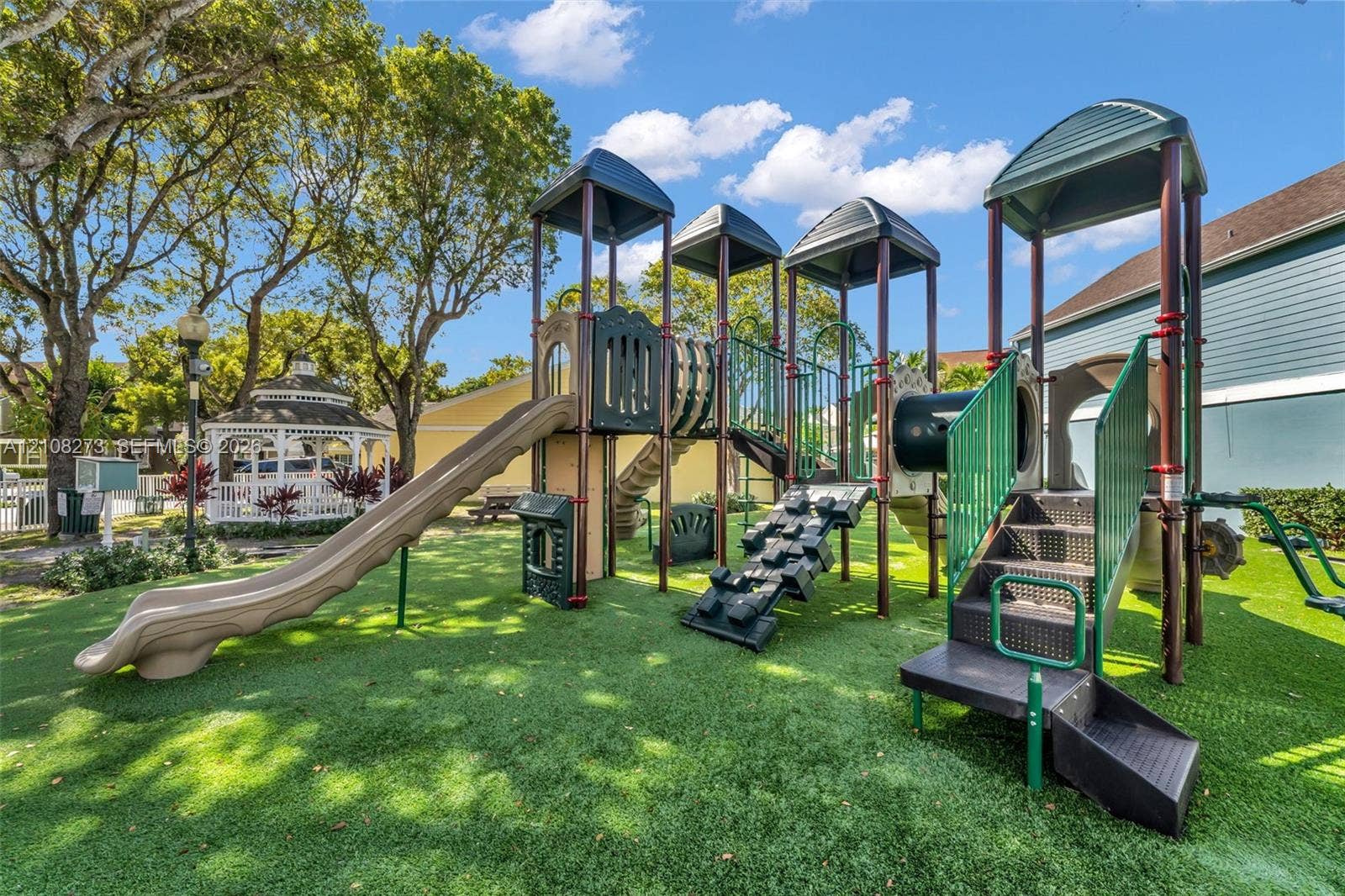 Playground area featuring multi-level climbing structures, dual slides, staircase access, and synthetic turf surfacing, situated on community grounds with a detached gazebo
