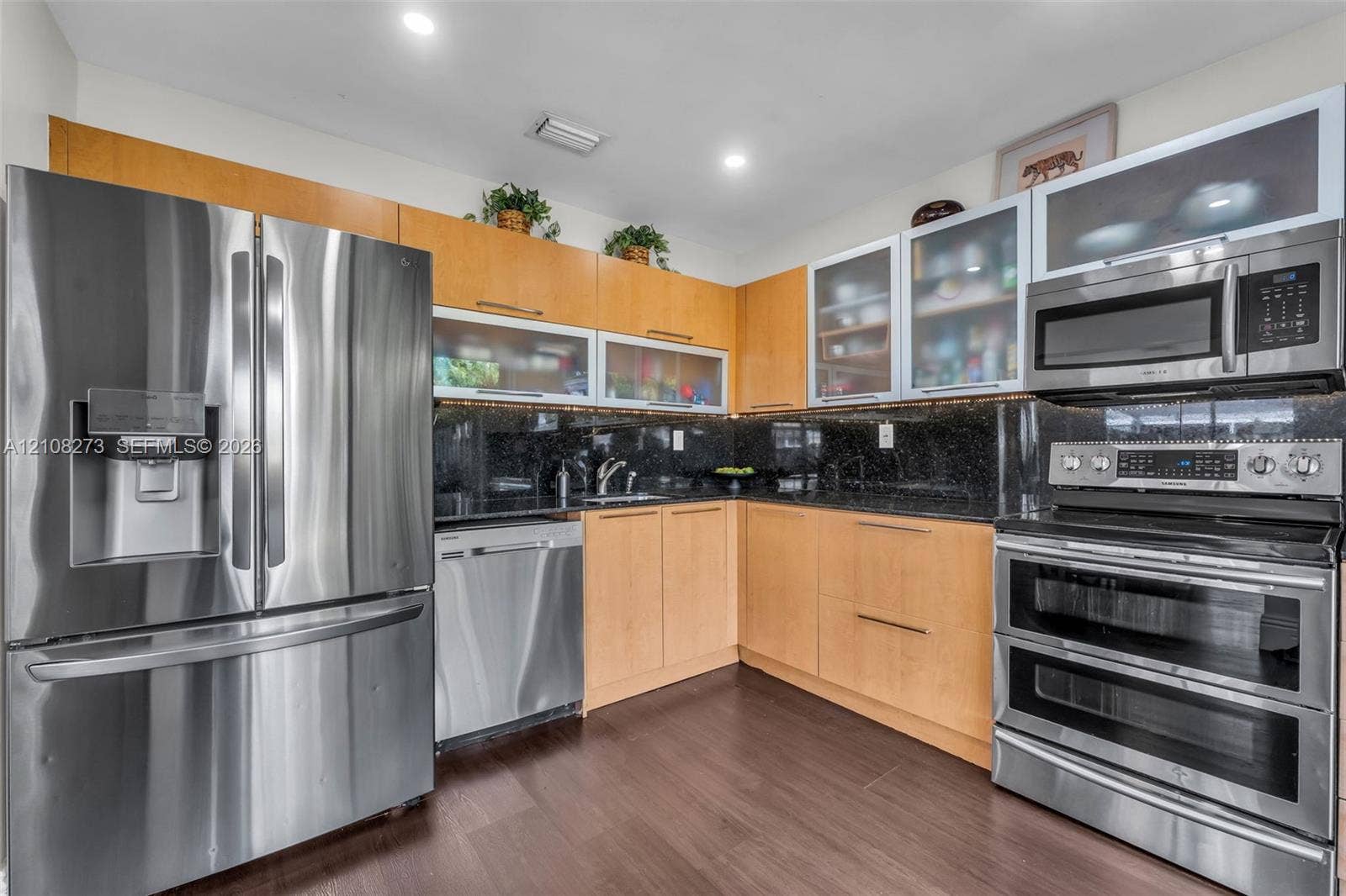 Kitchen featuring light wood-finish cabinetry with glass-front uppers, dark stone countertops, stainless steel appliances, and recessed ceiling lighting