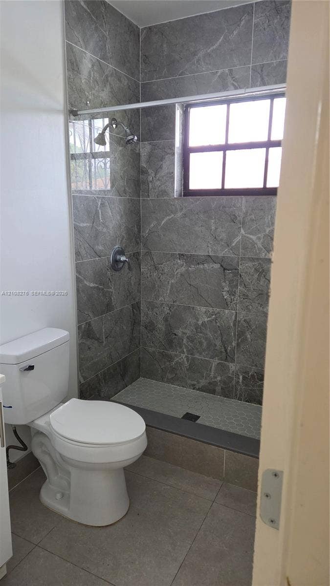 Walk-in shower featuring gray stone-finish tile surround, pebble-texture flooring, multi-pane window, and fixed chrome shower hardware