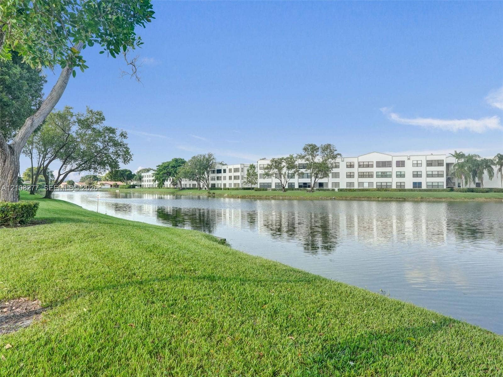 7466 Ashmont Circle, #303, Tamarac, FL 33321 Waterfront property featuring a manicured lawn, mature trees, and a lake shoreline